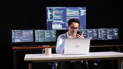 Programador masculino asiático que usa un teléfono inteligente mientras escribe código con una computadora portátil usando varios monitores