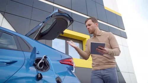 Man Charging Electric Car, Looks at Tablet in Frustration