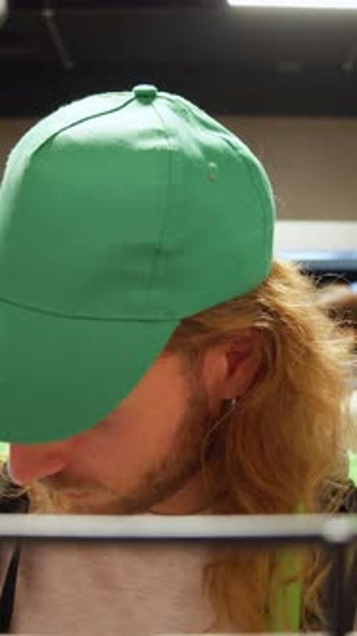 Face of Attractive Caucasian Man in Green Cap Courier Uniform Choosing Packs of Chips on Shelves