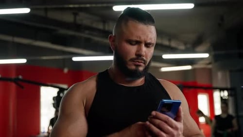 Muscular Man in Gym Using Smartphone Showing Focus and Contemplation During Workout Break