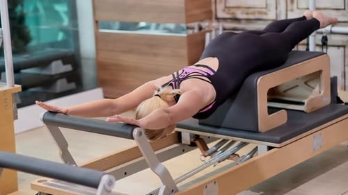 Pilates Practicing in Modern Gym Fit Blonde Coach Lying on Reformer Stretching Body