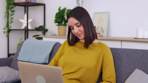 Woman Uses Laptop Smiling on Gray Couch Indoors
