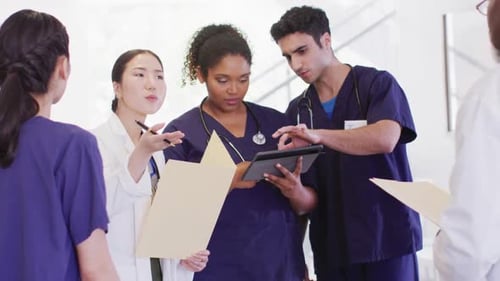 Video of diverse group of male and female doctors looking at tablet talking in hospital corridor