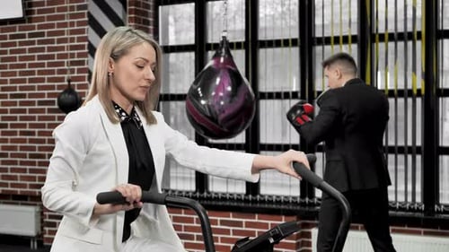 Couple of Businessmen Man and Woman Play Sports on an Exercise Bike and a Boxing Ring in Suits