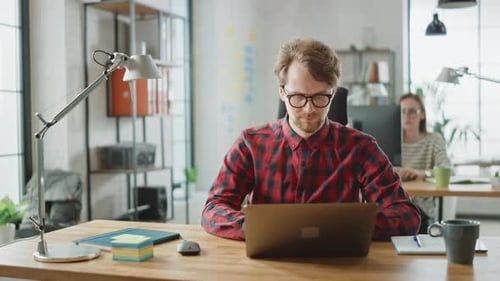 Handsome Young Man in Glasses and Shirt is Working on a Laptop in a Creative Business Agency. They
