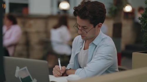 Focused Young Man Working Remotely on Laptop and Notebook in a Busy Cafe