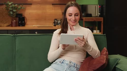 A Young Charming Smiling Caucasian Brunette in a Beige Blouse is Sitting on a Green Sofa in the