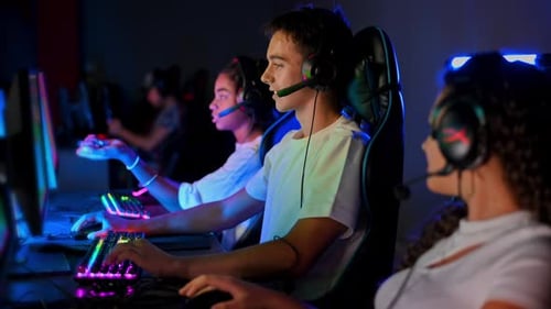 Multiracial group of teens in headsets playing video games in video game club with blue and red illu