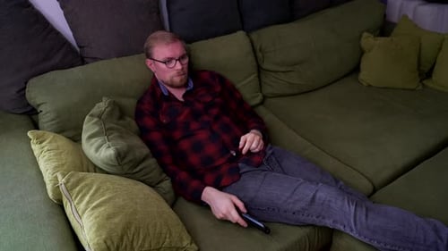 Young Man Lays on Sofa, Uses TV Remote to Switch Channels, Medium Shot