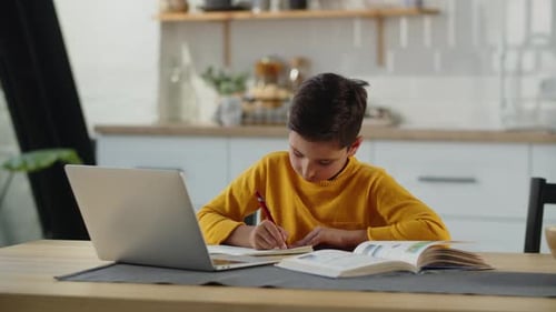 Der Junge sitzt an einem Tisch mit einem Buch und einem Laptop und schreibt etwas in ein Notizbuch Ein Junge in einem