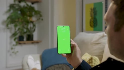 Man holding smartphone with green screen, touching it as if using it, sitting in the comfort of his