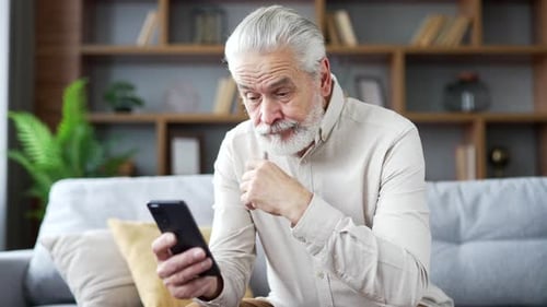 Mature Man Sitting on Couch Uses Smartphone