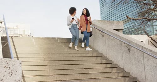 Diverse female friends descending plaza stairs responding to smartphone message and sipping coffee