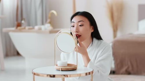 Young Beautiful Asian Woman Applying Red Lipstick on Lips Making Bright Makeup Sitting at Blurred