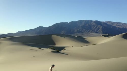 Drone footage of man walking through hot, dry sand dunes at sunset, in the desert, with mountains in