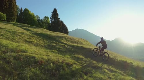 Drone is flying along an athletic man pedalling an MTB E-bike up a steep grassy hill.