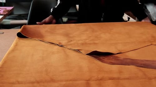 A man folding a large piece of leather on a workbench in a workshop where he created leather goods b