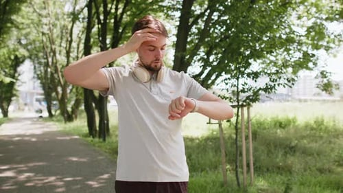 Active Young Man Jogger Running in Public Park and Looking at Smartwatch Getting Tired Relaxing