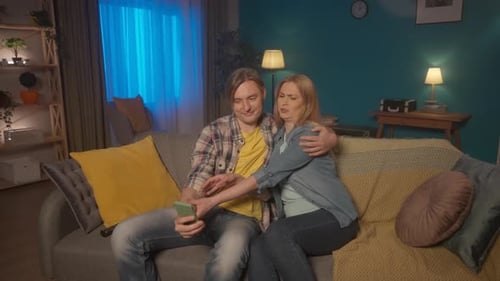 A Young Married Couple Sitting on the Couch at Home Makes a Joint Selfie The Couple Poses Hugging