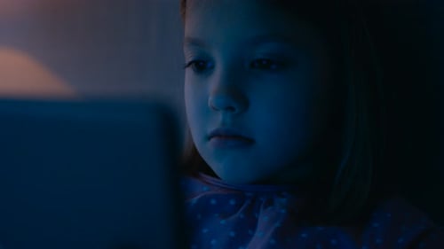 Süßes junges Mädchen liegt nachts in ihrem Bett und benutzt einen Tablet-Computer, der ihr Gesicht beleuchtet.
