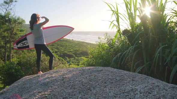 Woman surfer. Young woman stands with surfboard on the rock and checks ...