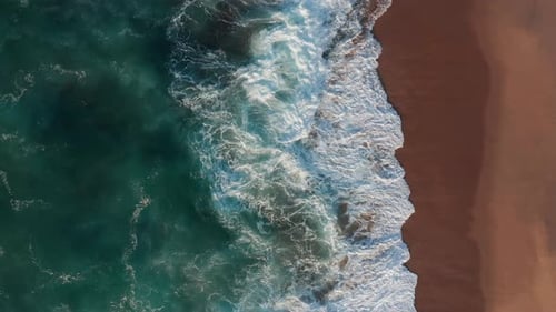 Aerial view tracking deserted beach with waves foaming over the sandy and rocky shoreline