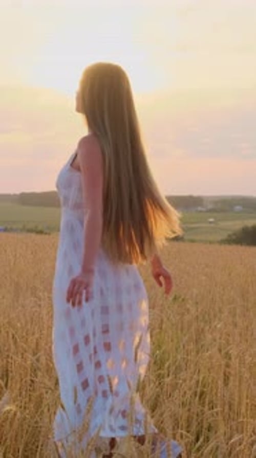 Woman with Long Hair in White Dress Walking Through Golden Wheat Field at Sunset with Soft Light