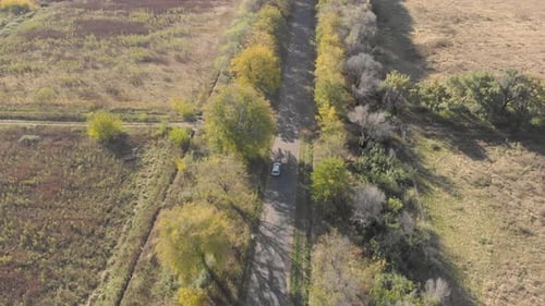 aerial following a grey car driving next to a green field on a tar road
