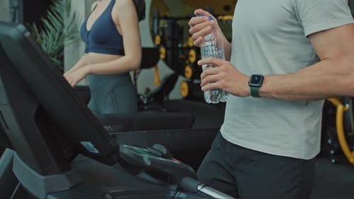 Man Drinking Water while Woman Works Out in Gym