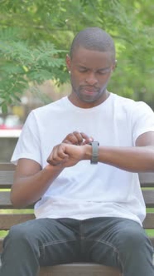 African American Man Using Smartwatch while Sitting on Bench in Park, Vertical Video