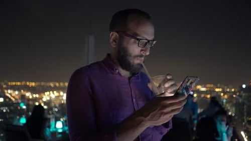 Handsome man texting on smartphone and sipping cocktail at rooftop bar at night