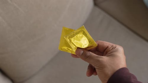 Hand Holding Two Condoms Indoors on Beige Couch