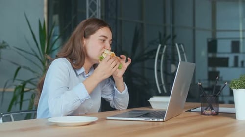 Young Businesswoman Eating Hamburger While Working on Laptop at the Office Table