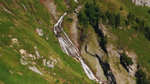 Aerial push in above alpine waterfall cascading on a steep, grassy and rocky mountainside on a sunny