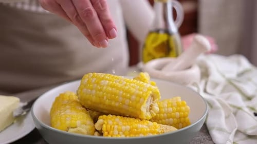 Woman Salting Freshly Cooked Corn in Ceramic Plate on a Kitchen Table