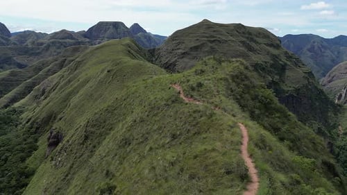 Aerial follows narrow grass ridge trail in rugged mountain landscape
