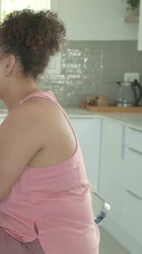 Vertical video: Shifting on stool, woman in pink tank testing lip balm at kitchen counter talking