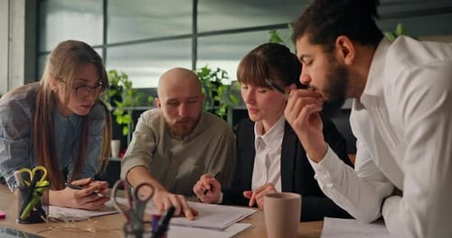 Diverse Team of Office Workers Gathers Around a Table Analyzing Data on Sheets of Paper and