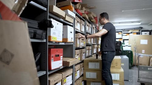 Homme européen avec des lunettes organisant des boîtes dans une salle de stockage remplie d'étagères et d'articles divers.