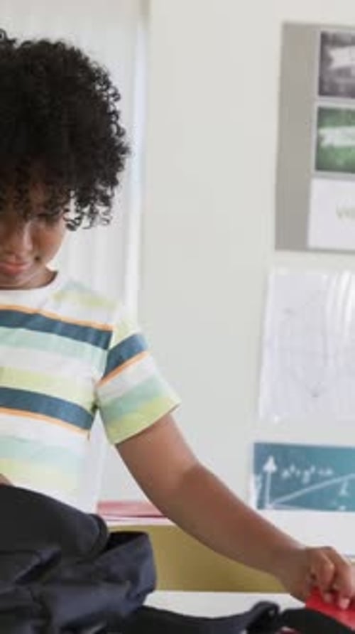 Vertical video: Packing kid in striped tee putting red book and ruler into black backpack at school