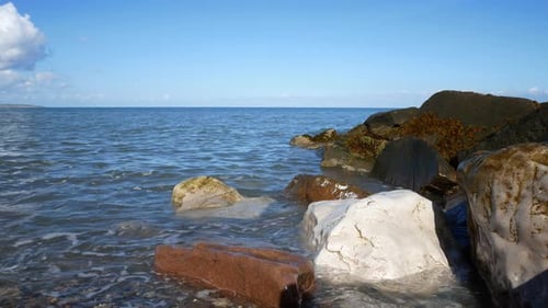 Sunny blue ocean tide washing up onto colourful stone pebble beach coastline island holiday scene do