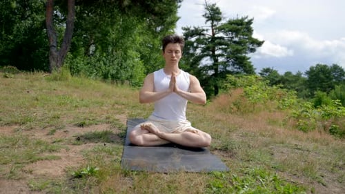 Man Sits in Lotus Pose on Mat on Meadow with Green Grass
