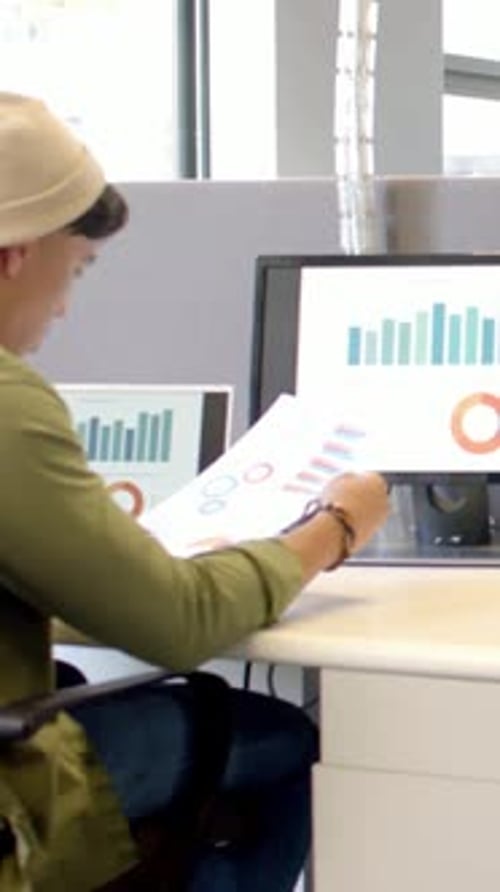 Vertical video: Comparing report man in olive top, hat checking data on screens at desk, copy space