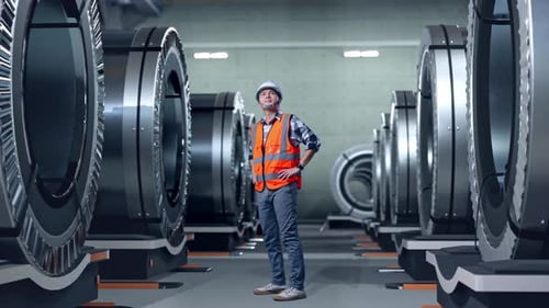 Side View Of Asian Male Engineer Looking Around With Arms Akimbo In Metal Factory