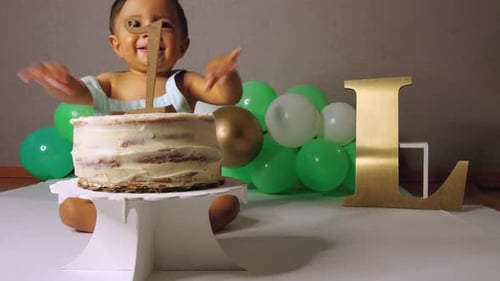 Cute latin baby toddler celebrating his birthday biting his cake and smiling with green balloons in
