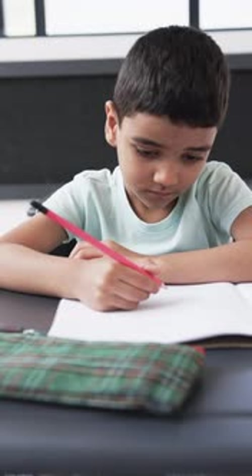 Vertical video: In a school setting, a young Caucasian boy focuses on writing