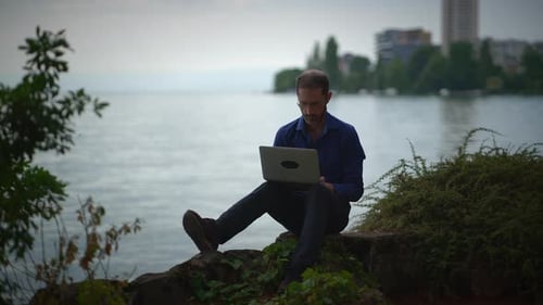 Man Working on Laptop by Lake in City