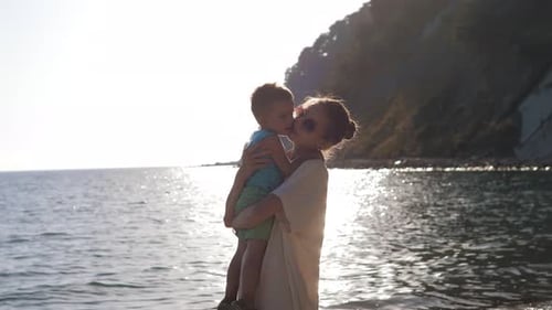 Young Mother of a 2 Years Old Boy is Keeping on Her Arms and Playing with a Son on a Seaside Beach