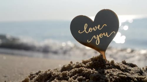 Herzförmiger Kunststoffstab mit Aufschrift „Ich liebe dich“, Hintergrund: Sand, Strand, Küste