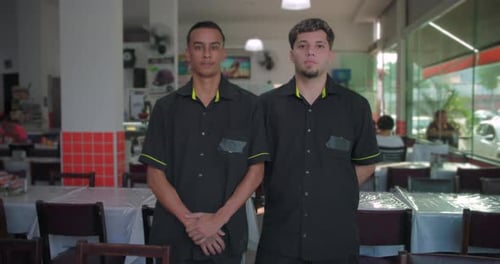 Two male restaurant employees standing side by side in dining area, hands folded in front, looking s